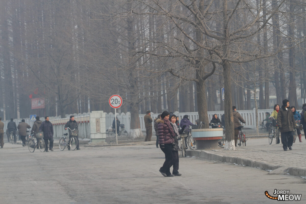 Everyday life scene in Pyongyang, North Korea: people walking, cyclists, urban setting, overcast sky.