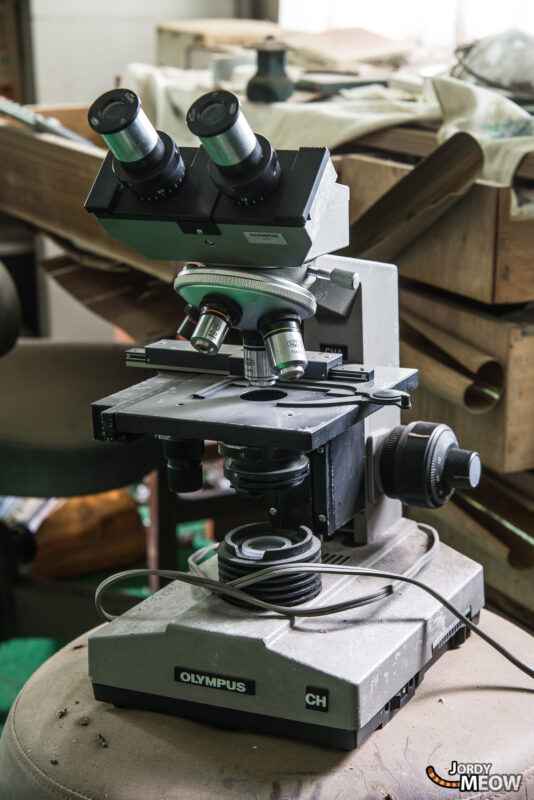 Dusty vintage microscope on cluttered bench inside an abandoned clinic hospital laboratory.