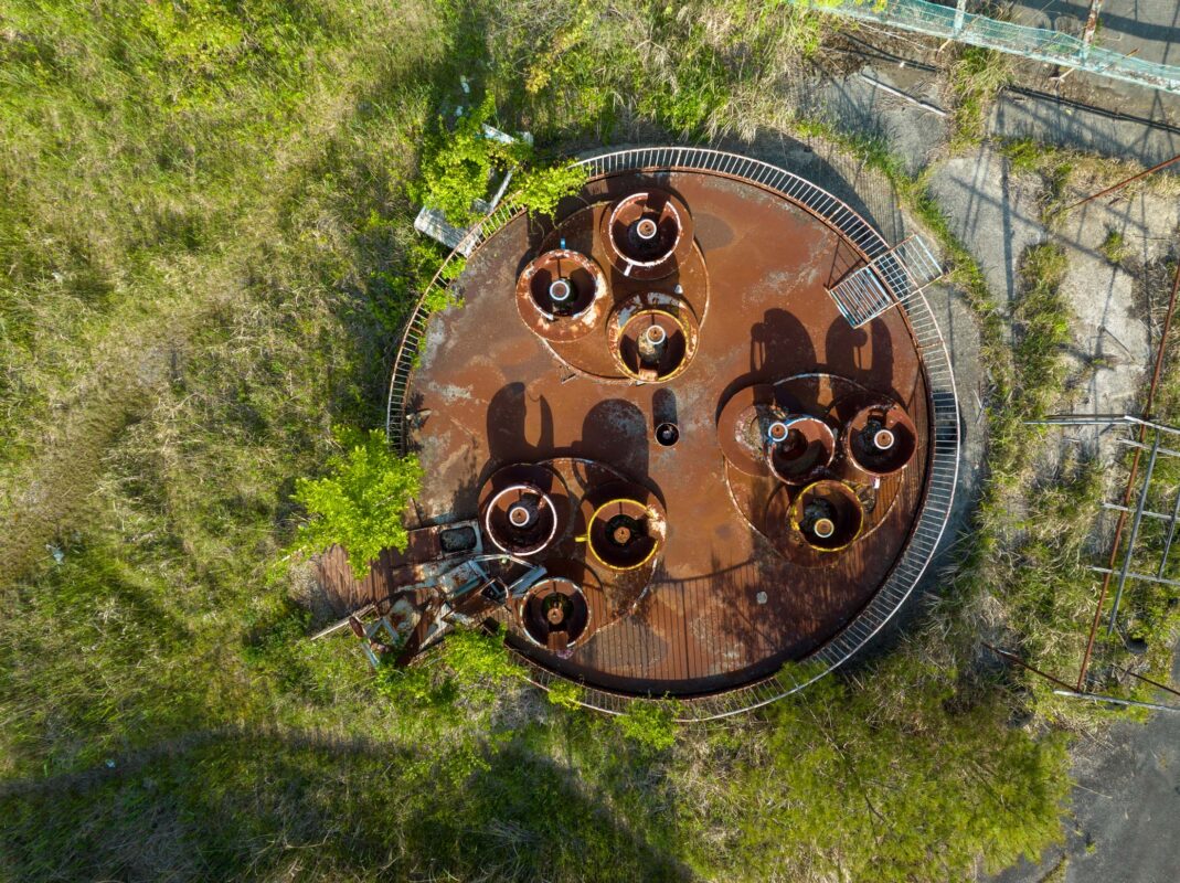 Aerial view of abandoned rusted amusement ride at Kejonuma Leisure Land, overgrown with grass.