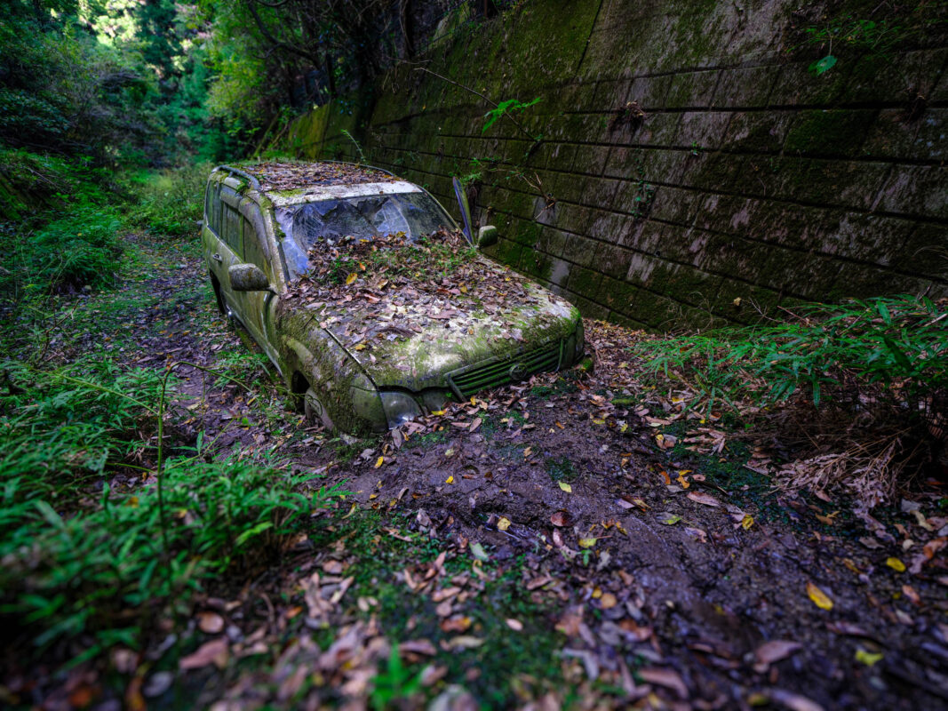 Abandoned Toyota Land Cruiser covered in moss, stuck in muddy forest beside stone wall