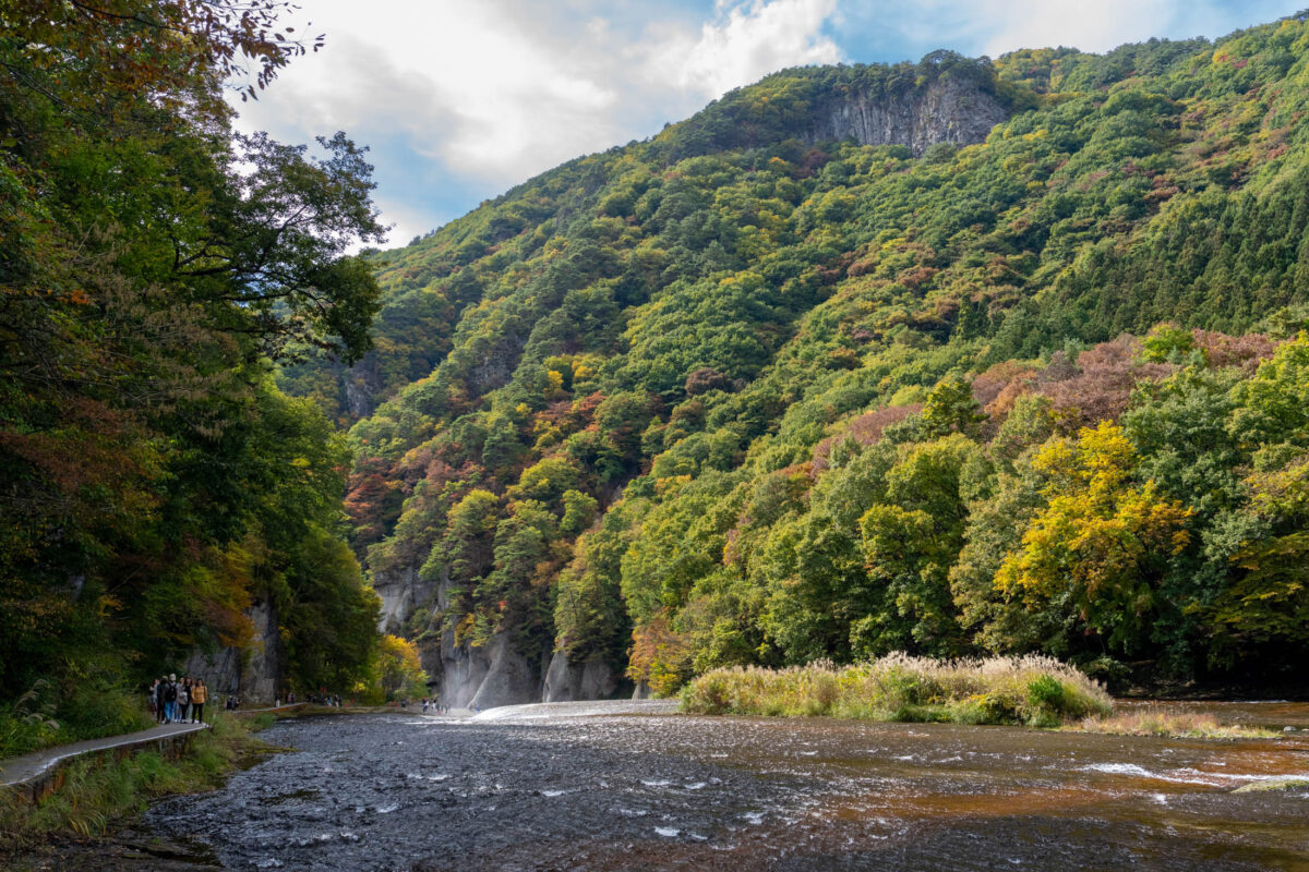 Fukiware Falls gorge with split cascade, autumn forest cliffs, and hikers on riverside trail.