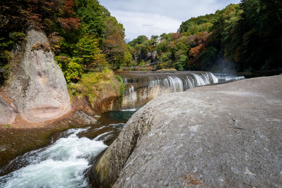 Fukiware Falls wide waterfall over rocky riverbed, surrounded by autumn forest in Japan.