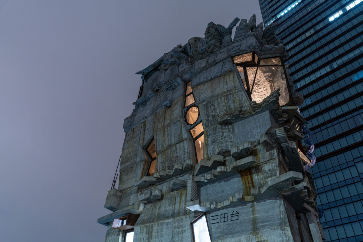 Weathered brutalist concrete landmark with glowing windows, set against a modern glass high-rise.