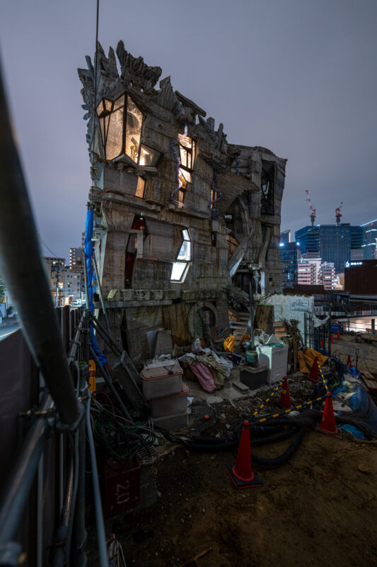 Warped partially demolished building glowing at dusk, construction debris below, modern skyline behind.
