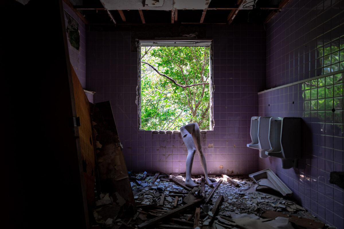 Mannequin legs in abandoned resort restroom with purple tiles, urinals, and overgrown window.
