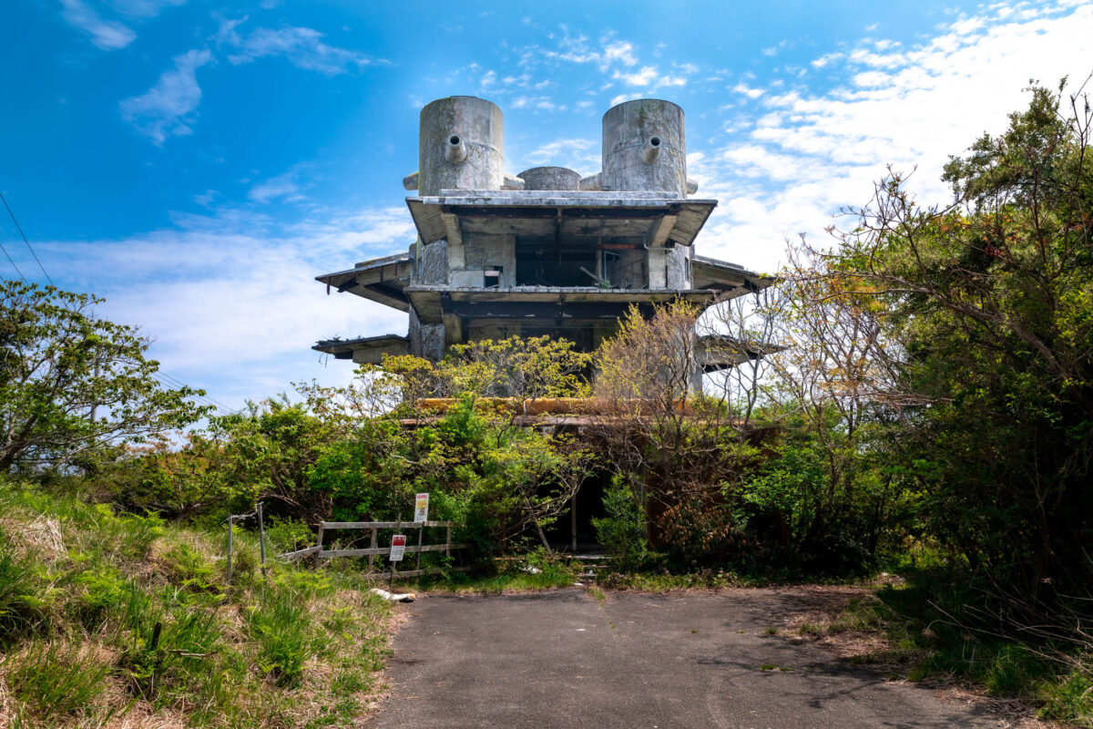 Abandoned New Muroto Sky Resort, brutalist concrete towers above overgrown path and greenery.