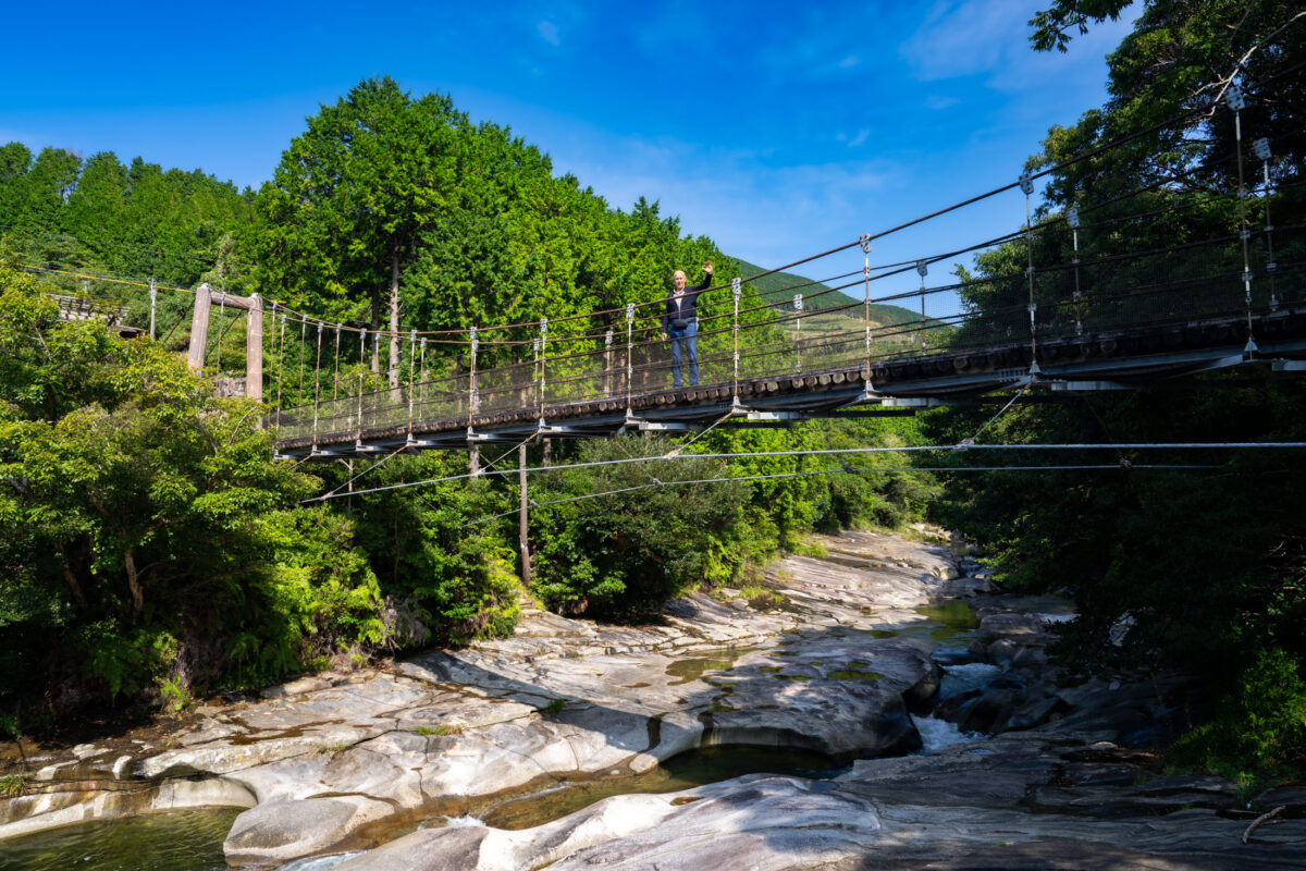 Suspension bridge over Ayumodoshi Natural Park river gorge, surrounded by lush green forest.