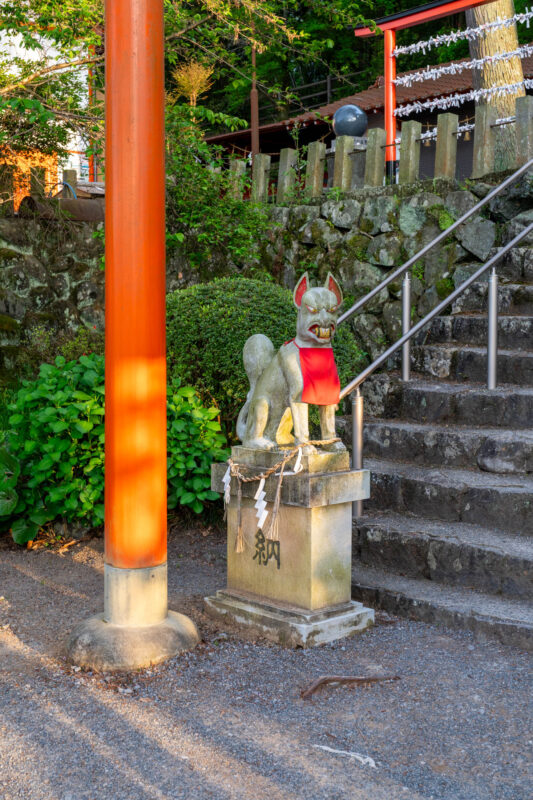 Stone fox guardian with red bib beside vermilion torii at Ukiha Inari Shrine staircase.