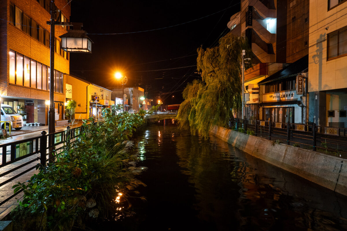 Izuhara canal at night with warm streetlights, building reflections, and willow tree