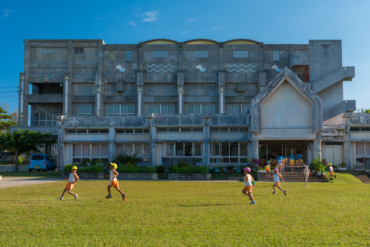 Children in sun hats running across grassy field by concrete school, Miyako Island, Japan.