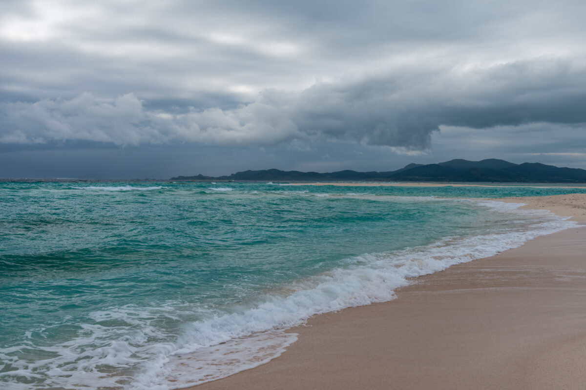 Kumejima beach with turquoise waves, distant hills, and stormy clouds overhead