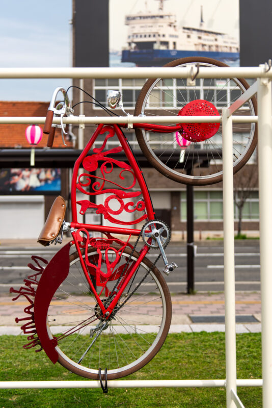 Upside-down red bicycle sculpture hanging at Naoshima Harbor with ferry in background.
