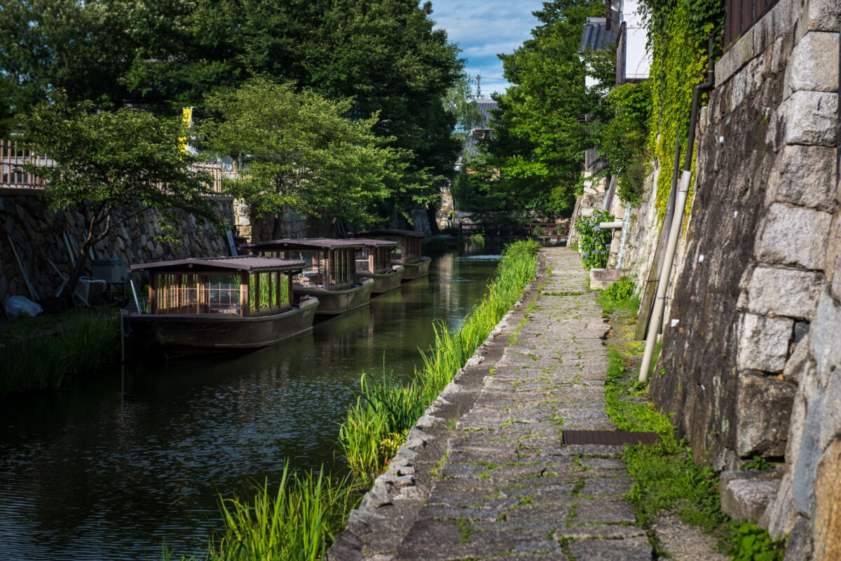Omihachiman canal in Japan with traditional wooden boats and stone walkway under trees.