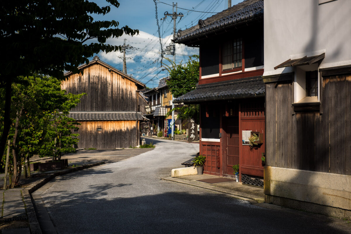 Sunlit historic street in Omihachiman, Shiga, Japan with traditional wooden buildings and tiled roofs.
