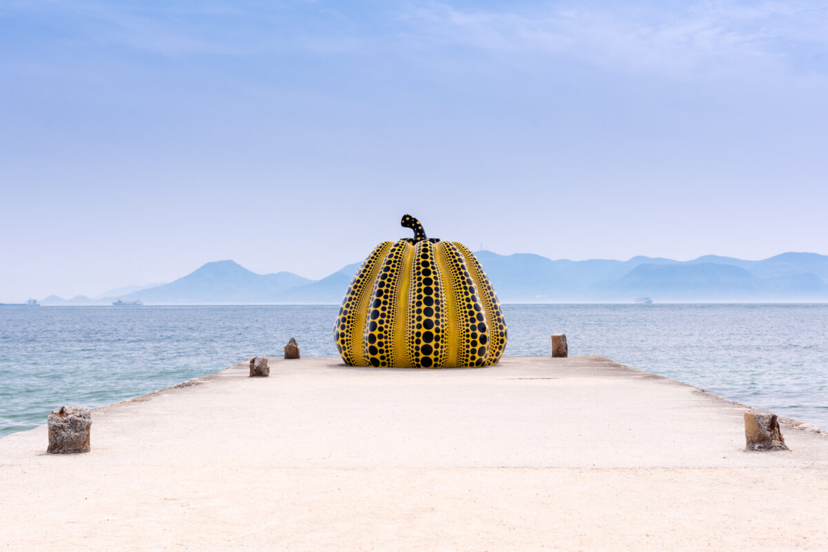 Yellow polka-dot pumpkin sculpture on Naoshima pier overlooking calm sea and distant mountains.