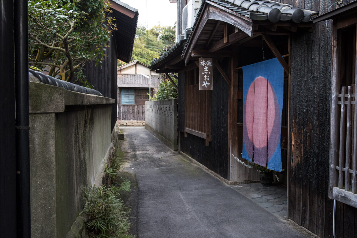 Quiet Naoshima Island alley with traditional wooden houses and blue noren curtain.