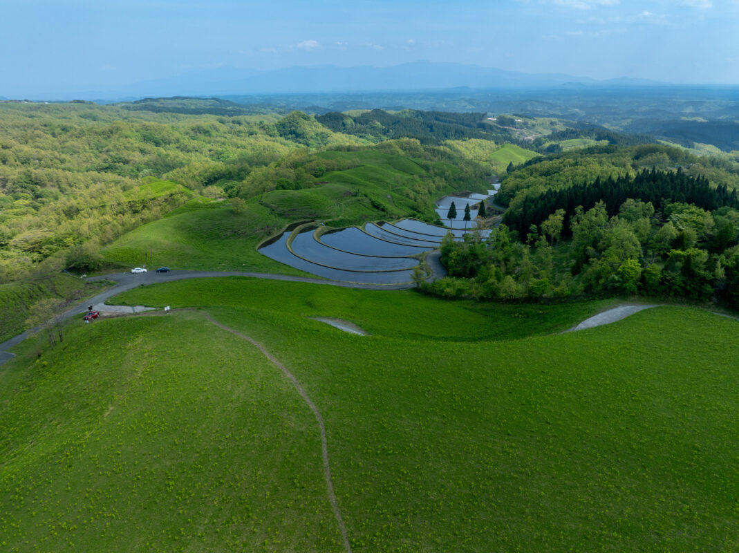 Aerial view of Ogi terraced rice paddies, curved water-filled fields, winding road, and distant hills.